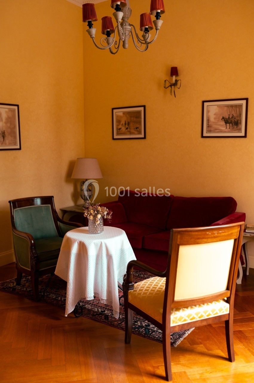 Salon chaleureux avec canapé rouge, deux fauteuils, table ronde recouverte d'une nappe blanche et décorations murales.