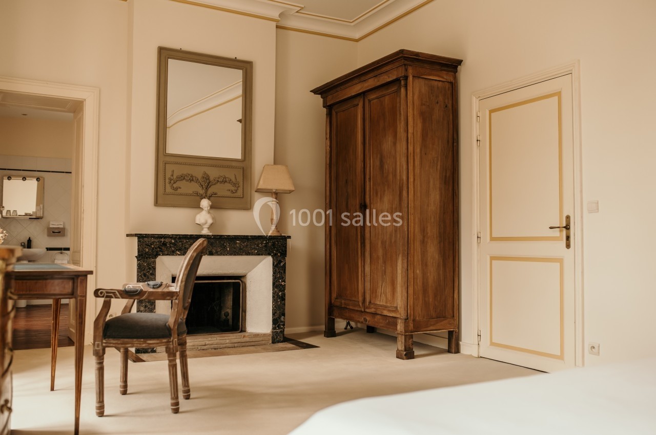 Chambre lumineuse avec armoire en bois, cheminée décorative, bureau, chaise et miroir au mur.