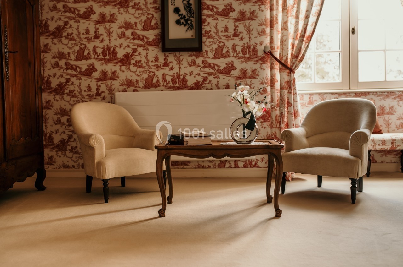 Deux fauteuils beiges entourent une table basse en bois avec un vase de fleurs, dans une pièce au décor classique.