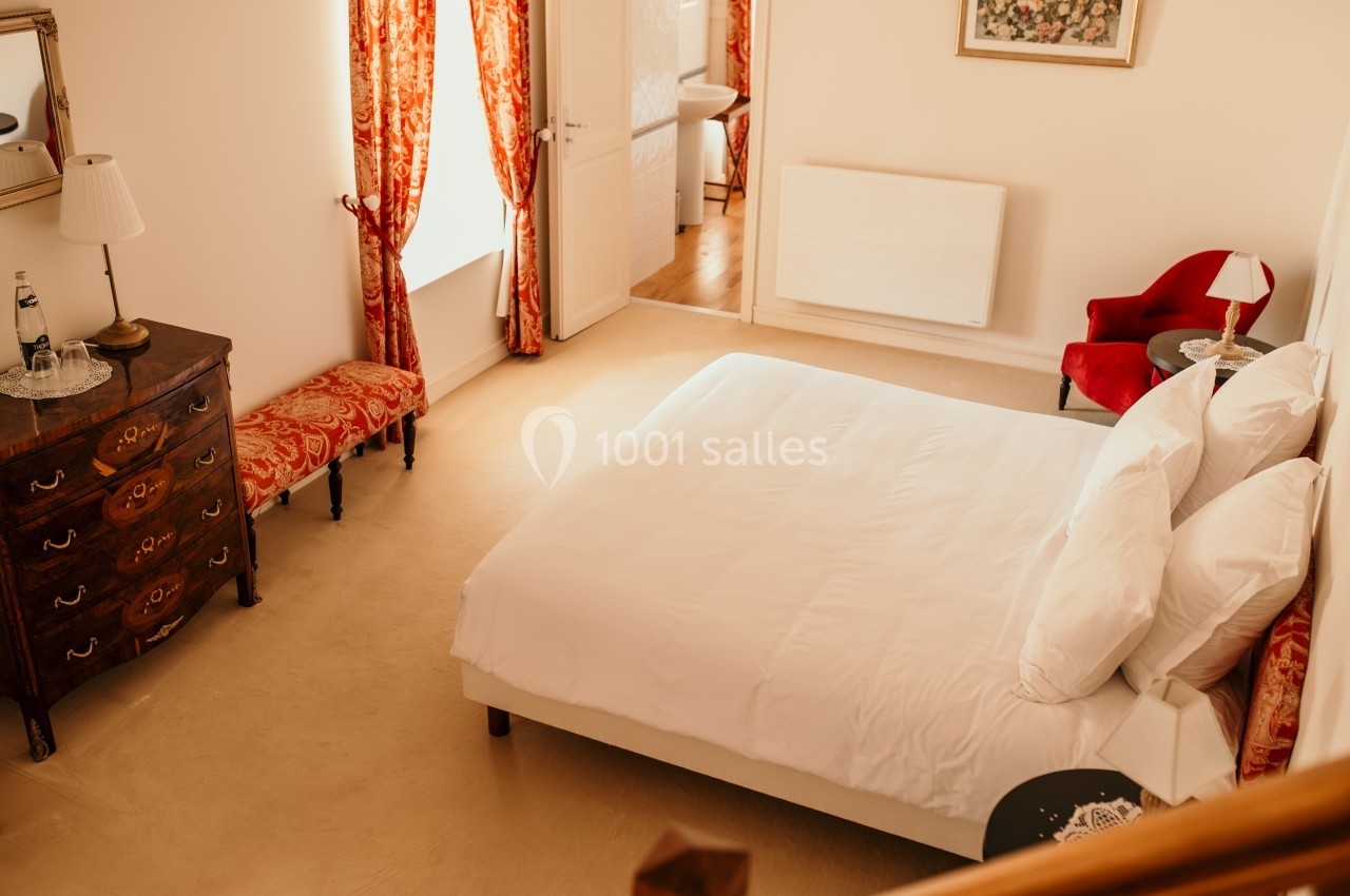 Chambre lumineuse avec lit double, draps blancs, fauteuil rouge, commode en bois et rideaux rouges à motifs.