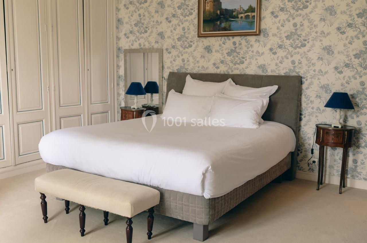 Chambre avec un lit double, tête de lit grise, tables de chevet en bois et mur décoré de papier peint fleuri.