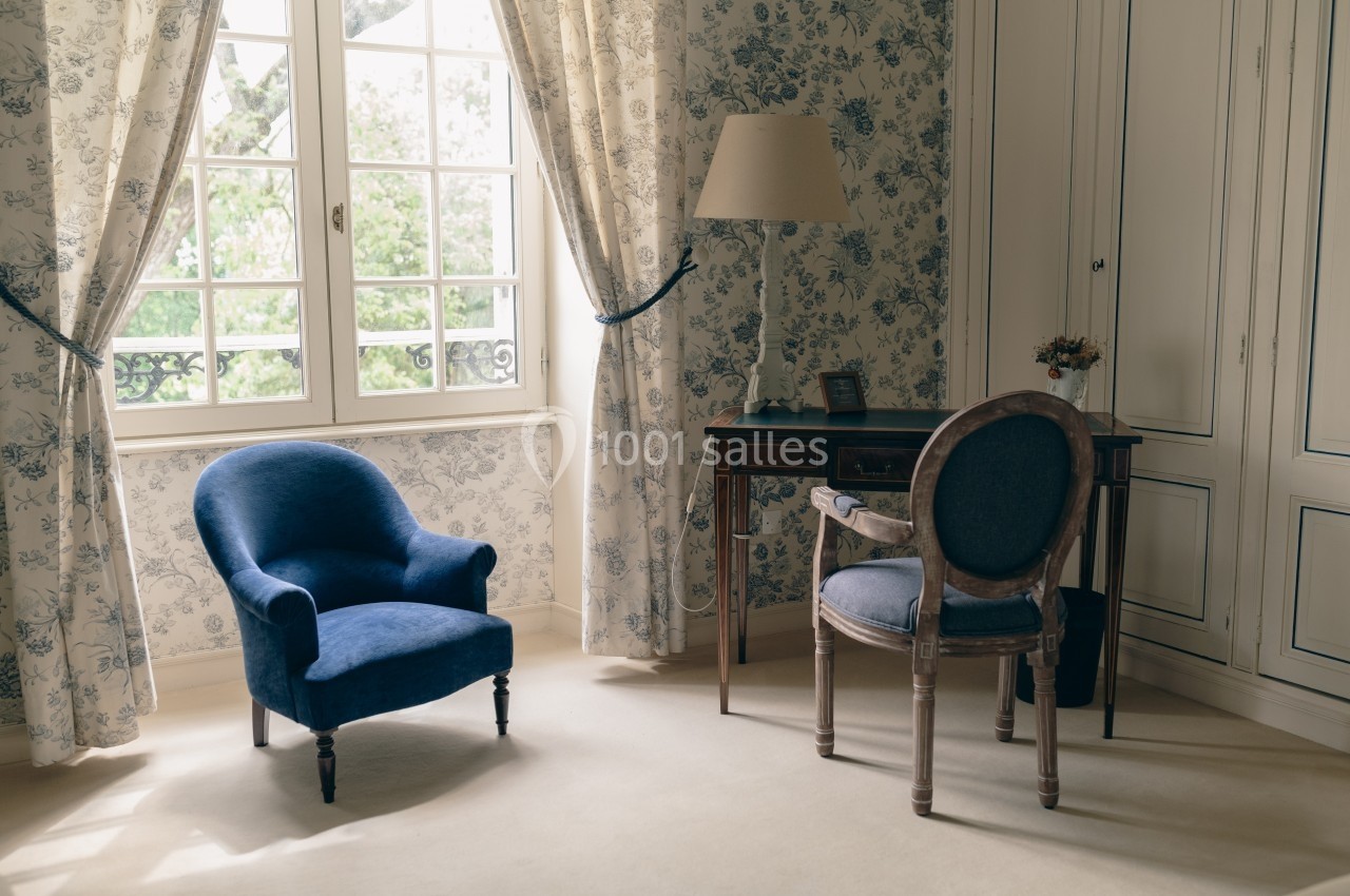 Fauteuil bleu, bureau en bois et chaise dans une pièce lumineuse avec papier peint fleuri et grandes fenêtres.