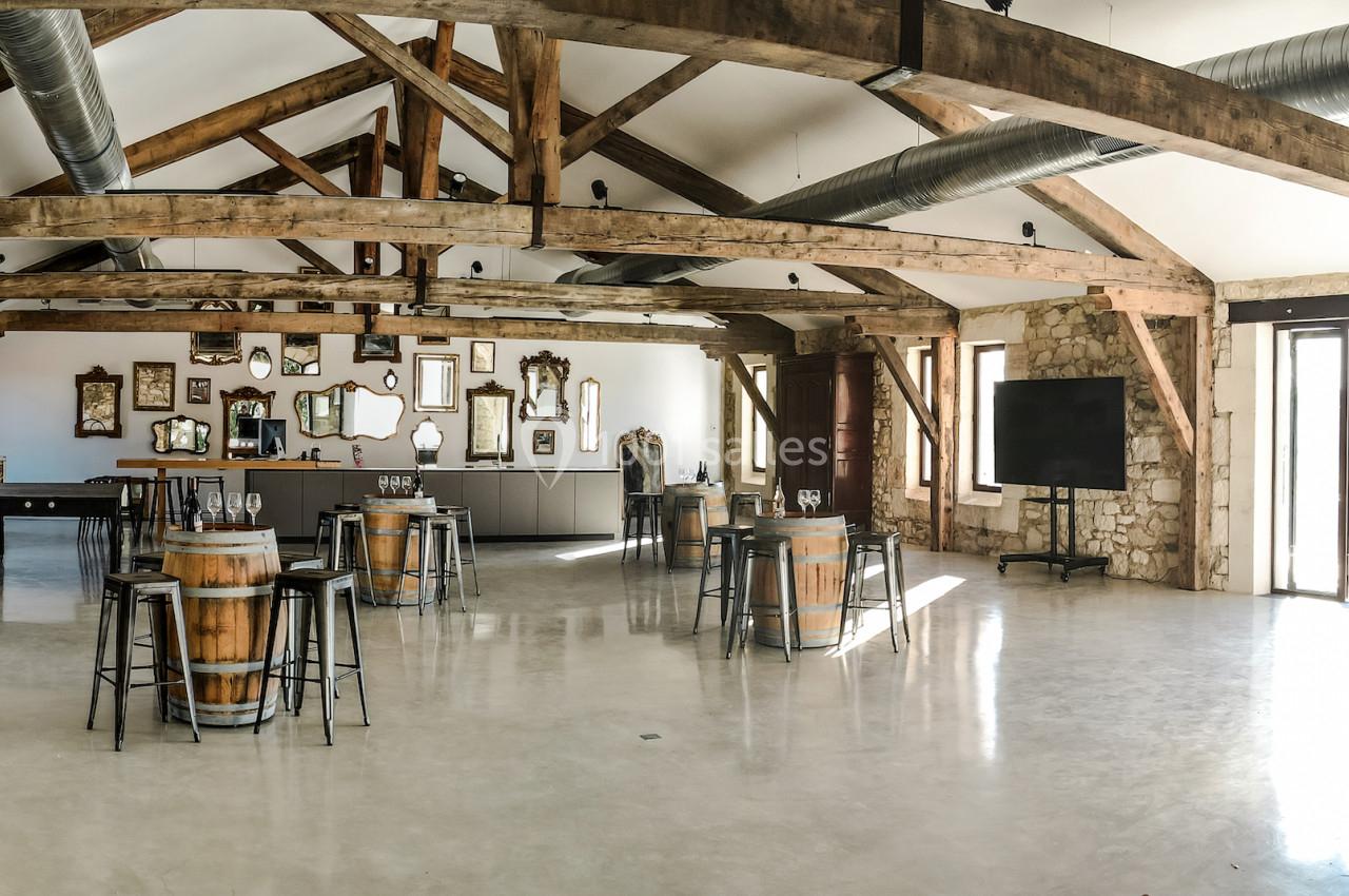 Salle lumineuse avec poutres apparentes, tables en tonneaux, chaises hautes, murs en pierre et vue sur l'extérieur.