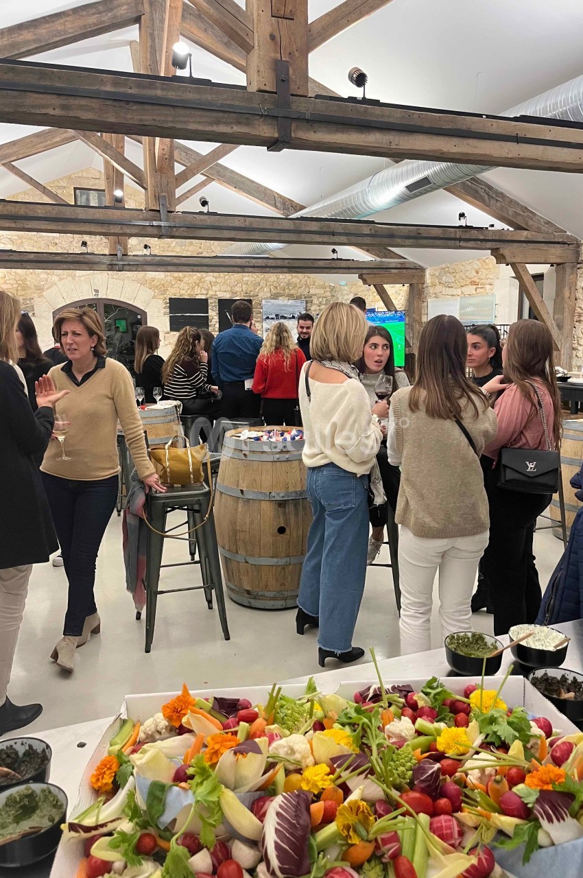 Des personnes discutent dans une salle lumineuse avec des poutres apparentes, un buffet de légumes colorés au premier plan.
