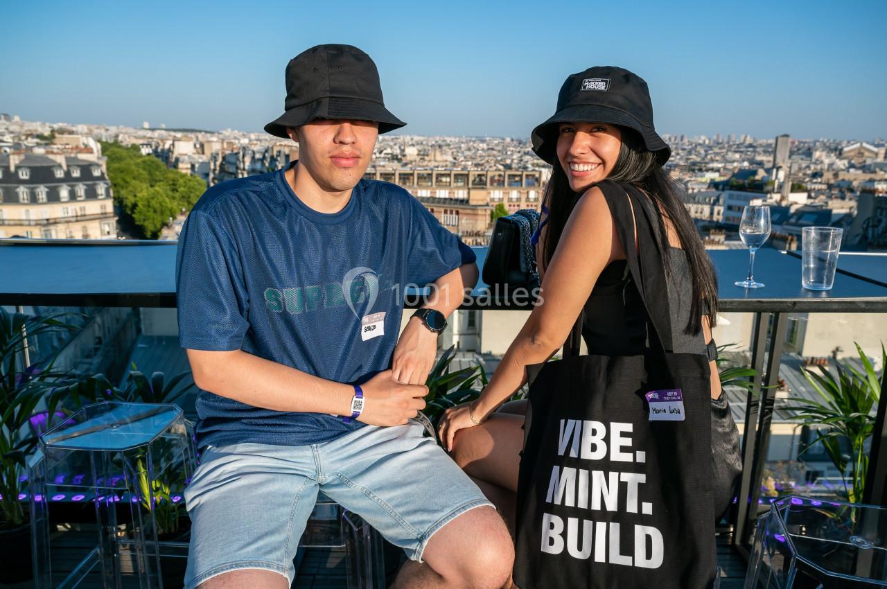 Deux personnes assises sur une terrasse ensoleillée avec vue sur des bâtiments urbains, portant des chapeaux noirs.