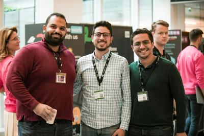 Trois hommes souriants posent ensemble dans un espace professionnel lors d'un événement, badges autour du cou.