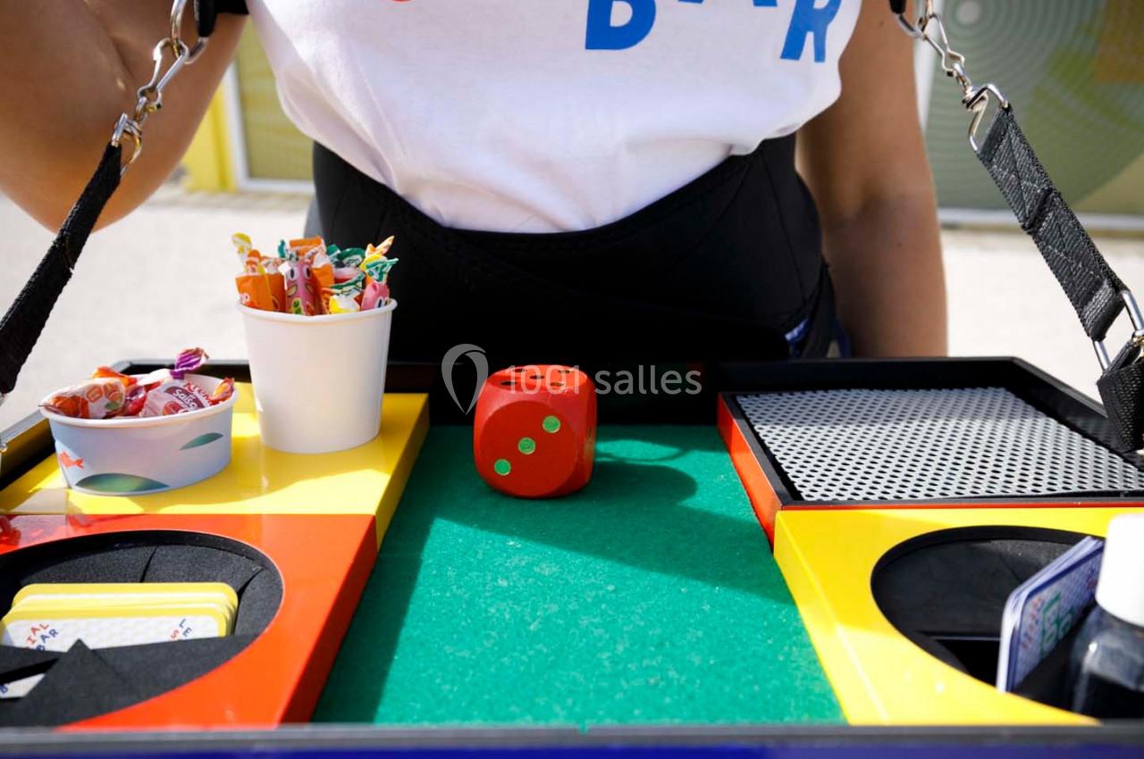 Plateau coloré contenant un dé rouge, des cartes, des bonbons et divers accessoires, tenu par une personne.