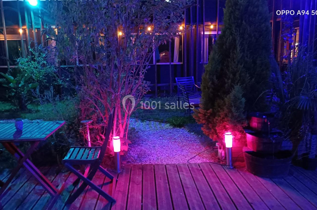Terrasse en bois éclairée par des lumières colorées, entourée de végétation et de mobilier de jardin.