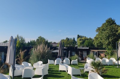 Terrain de pétanque en gravier entouré de verdure, avec des tables et une tente blanche en arrière-plan.