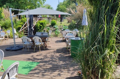 Terrain de pétanque en gravier entouré de verdure, avec des tables et une tente blanche en arrière-plan.