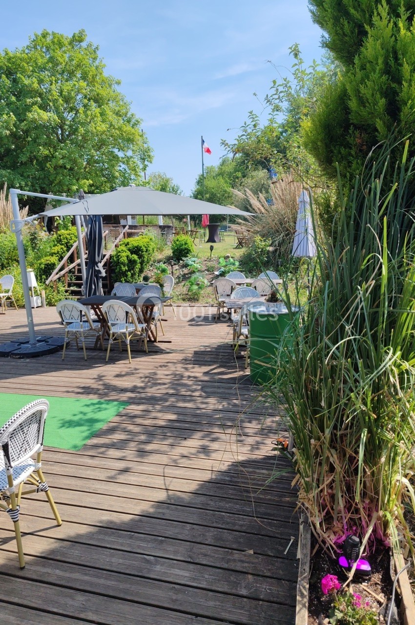 Terrasse en bois avec tables et chaises, entourée de végétation, sous un ciel bleu et un drapeau visible au loin.