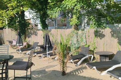 Terrain de pétanque en gravier entouré de verdure, avec des tables et une tente blanche en arrière-plan.