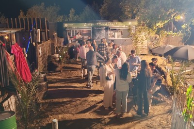 Terrain de pétanque en gravier entouré de verdure, avec des tables et une tente blanche en arrière-plan.