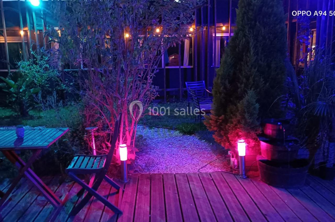 Location salle Hem (Nord) - O'Green du Château d'Hem #9 Terrasse en bois éclairée par des lumières colorées, entourée de végétation et de mobilier de jardin.