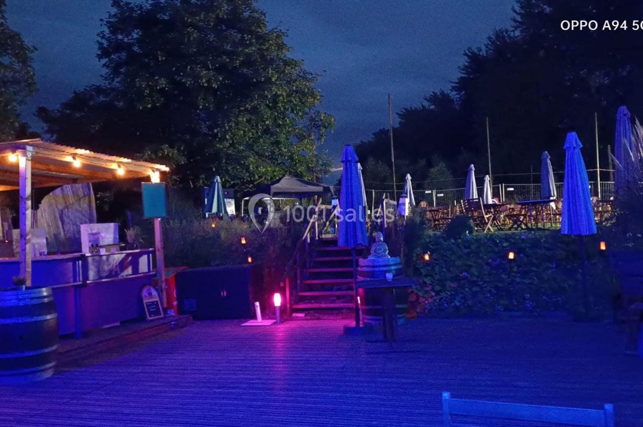 Location salle Hem (Nord) - O'Green du Château d'Hem #18 Terrasse extérieure éclairée en soirée avec des parasols, des tables et un bar en bois sous des lumières tamisées.