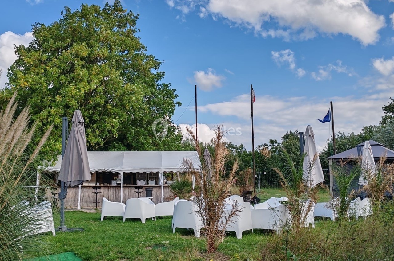 Location salle Hem (Nord) - O'Green du Château d'Hem #26 Espace extérieur avec chaises blanches, parasols fermés, tente et drapeaux, entouré de végétation sous un ciel bleu.