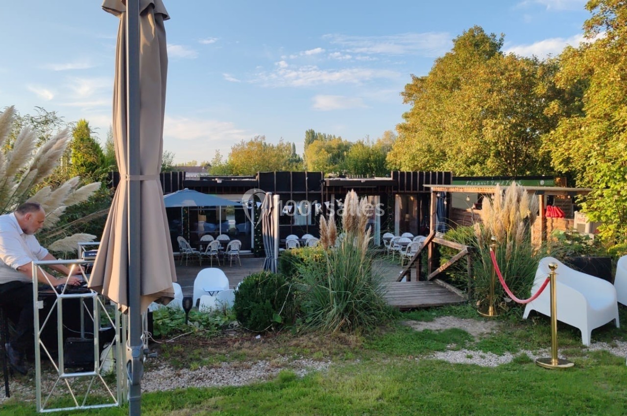 Location salle Hem (Nord) - O'Green du Château d'Hem #30 Un homme assis à une table en extérieur près d'une terrasse entourée de végétation et de mobilier de jardin.