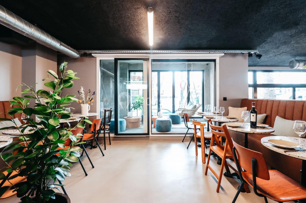 Salle de restaurant moderne avec tables dressées, chaises en bois, plantes décoratives et grande baie vitrée.