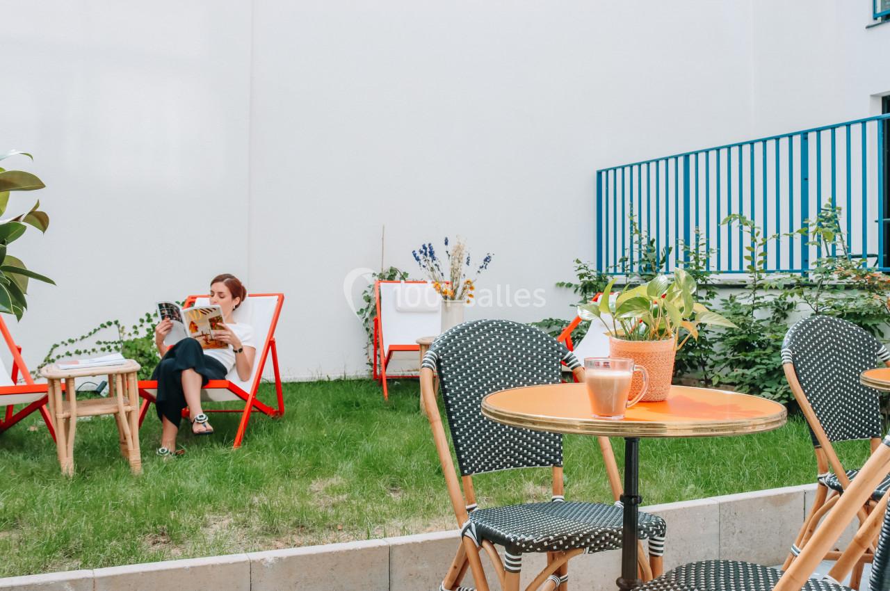 Une femme lit sur une chaise longue dans un jardin, entourée de plantes et de mobilier de terrasse coloré.