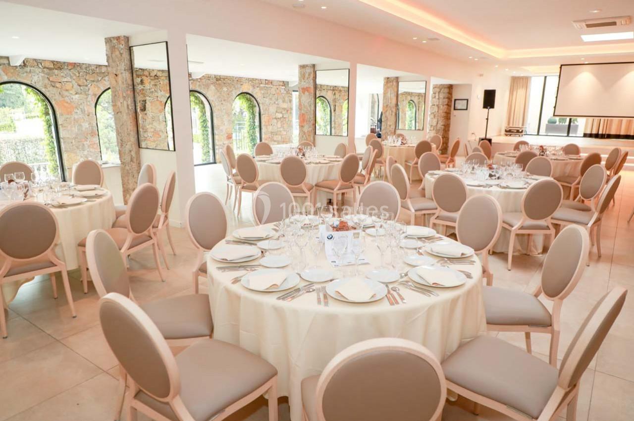 Salle de réception lumineuse avec tables rondes dressées, chaises beige clair et murs en pierre apparente.