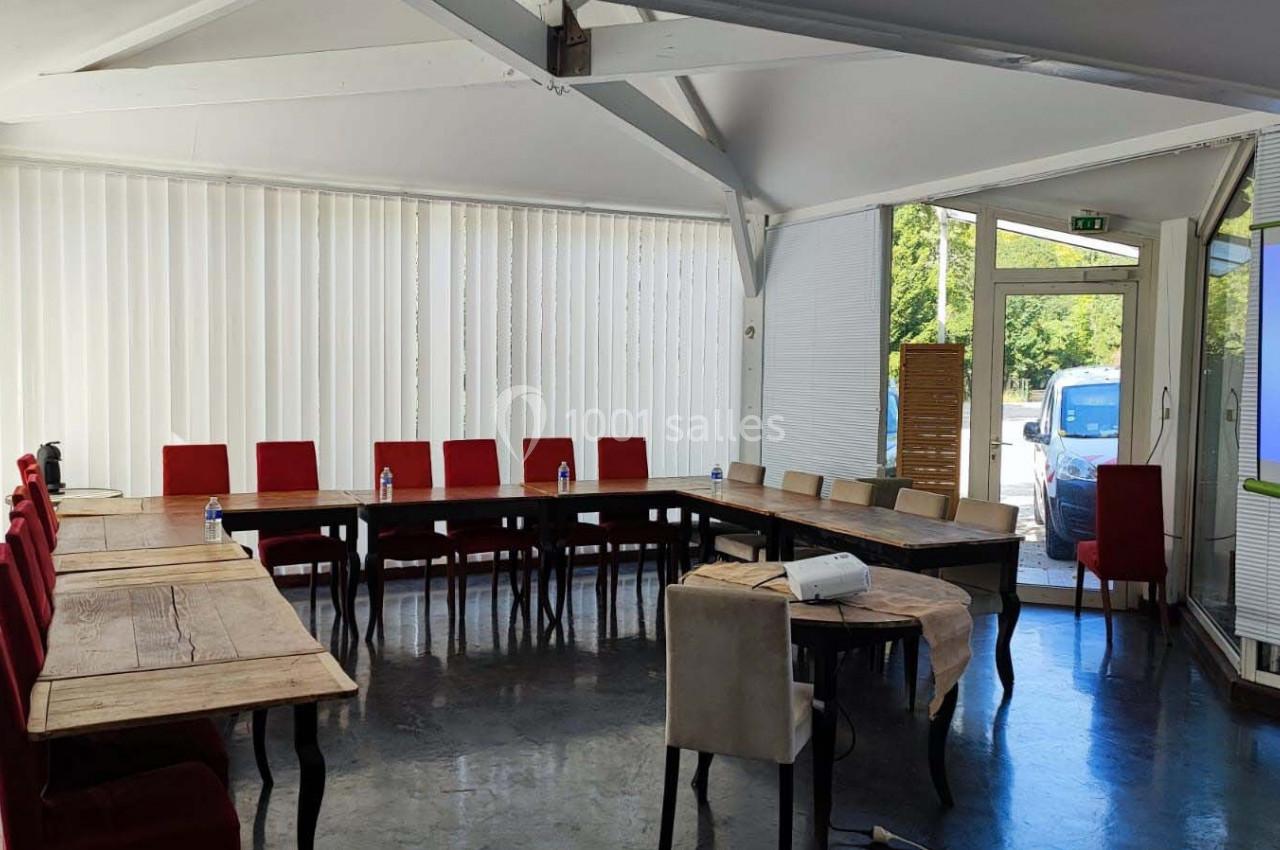 Salle de réunion lumineuse avec des tables en bois disposées en U, des chaises rouges et un projecteur allumé.