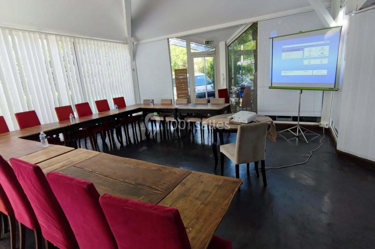 Salle de réunion lumineuse avec tables en bois disposées en U, chaises rouges et écran de projection allumé.