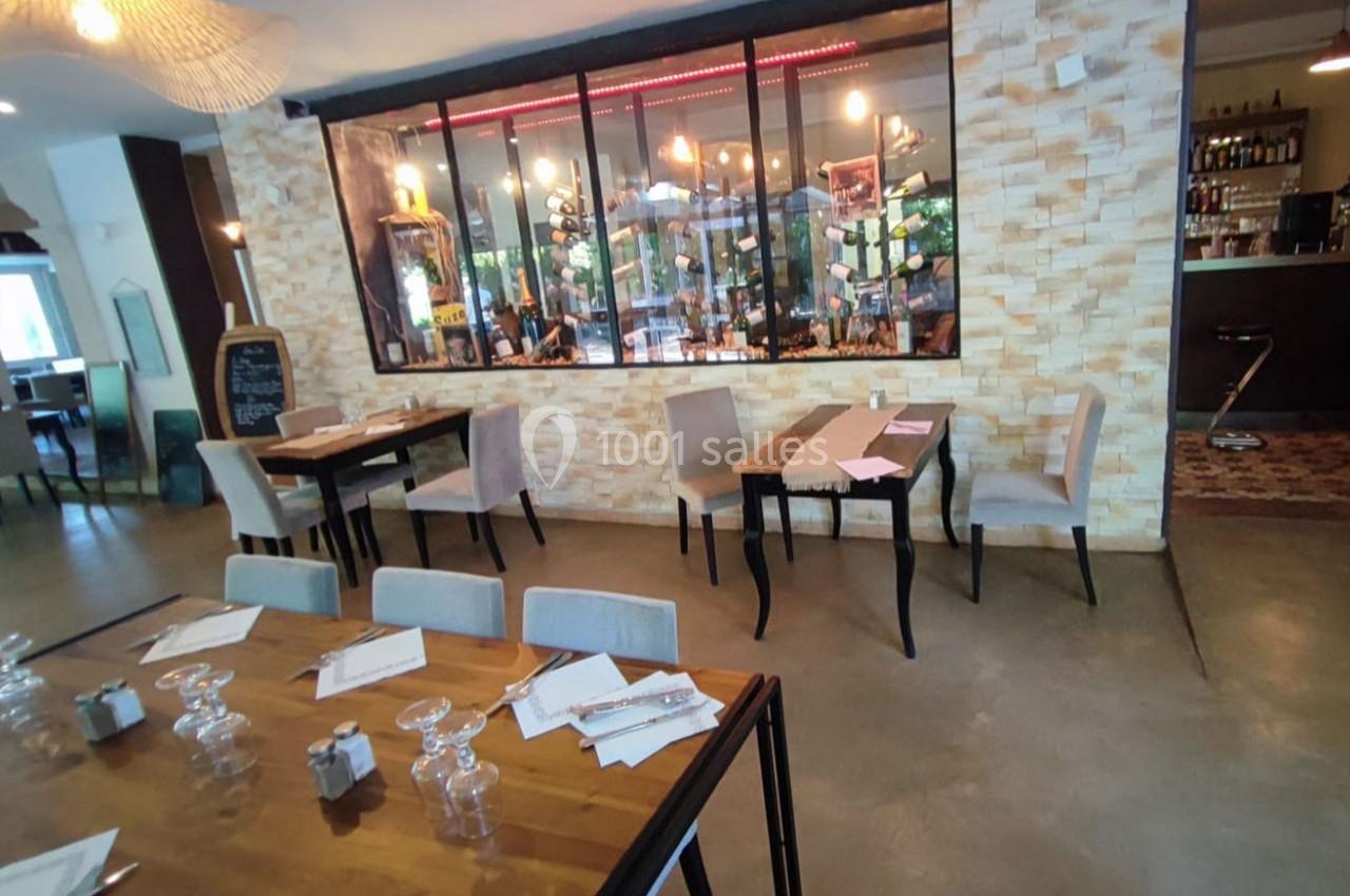 Salle de restaurant moderne avec tables en bois, chaises rembourrées, et vue sur une cave à vin derrière une vitre.