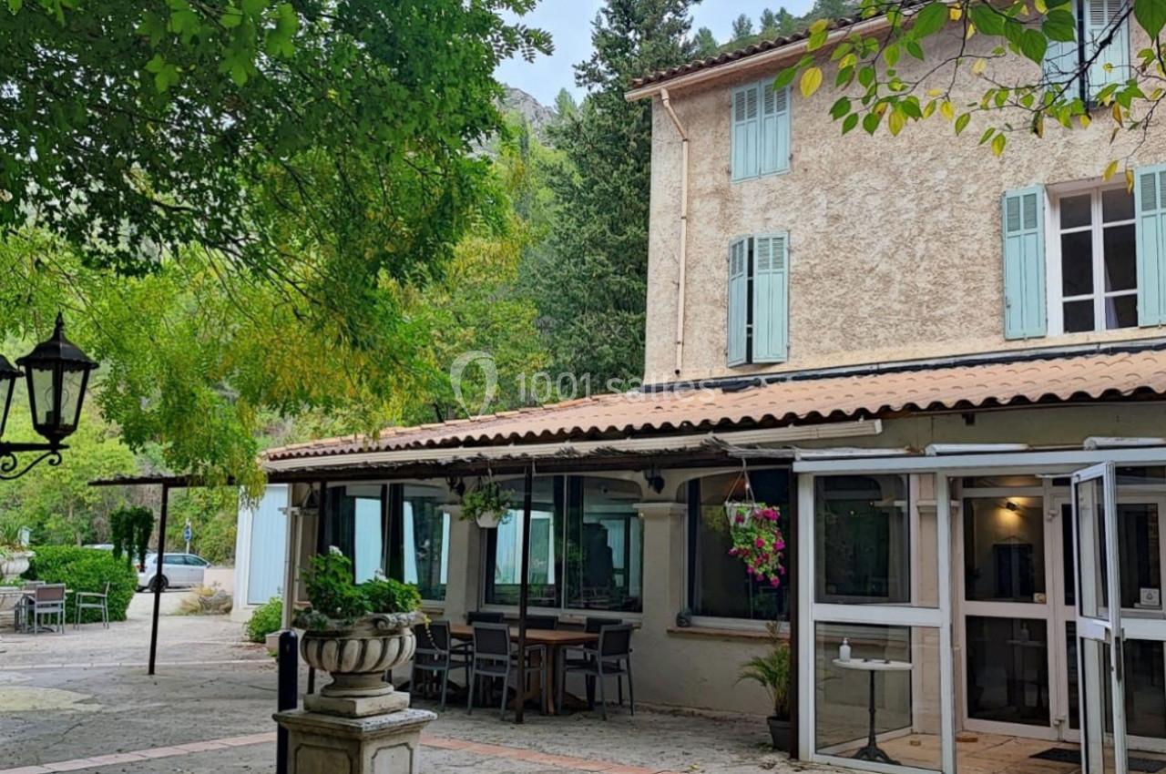 Façade d'une maison en pierre avec volets bleus, terrasse aménagée et végétation environnante.