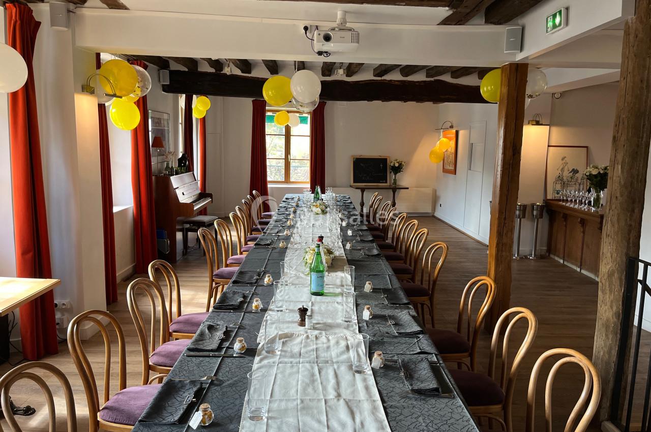 Salle décorée avec une longue table dressée, chaises en bois, ballons jaunes et lumière naturelle entrant par les fenêtres.