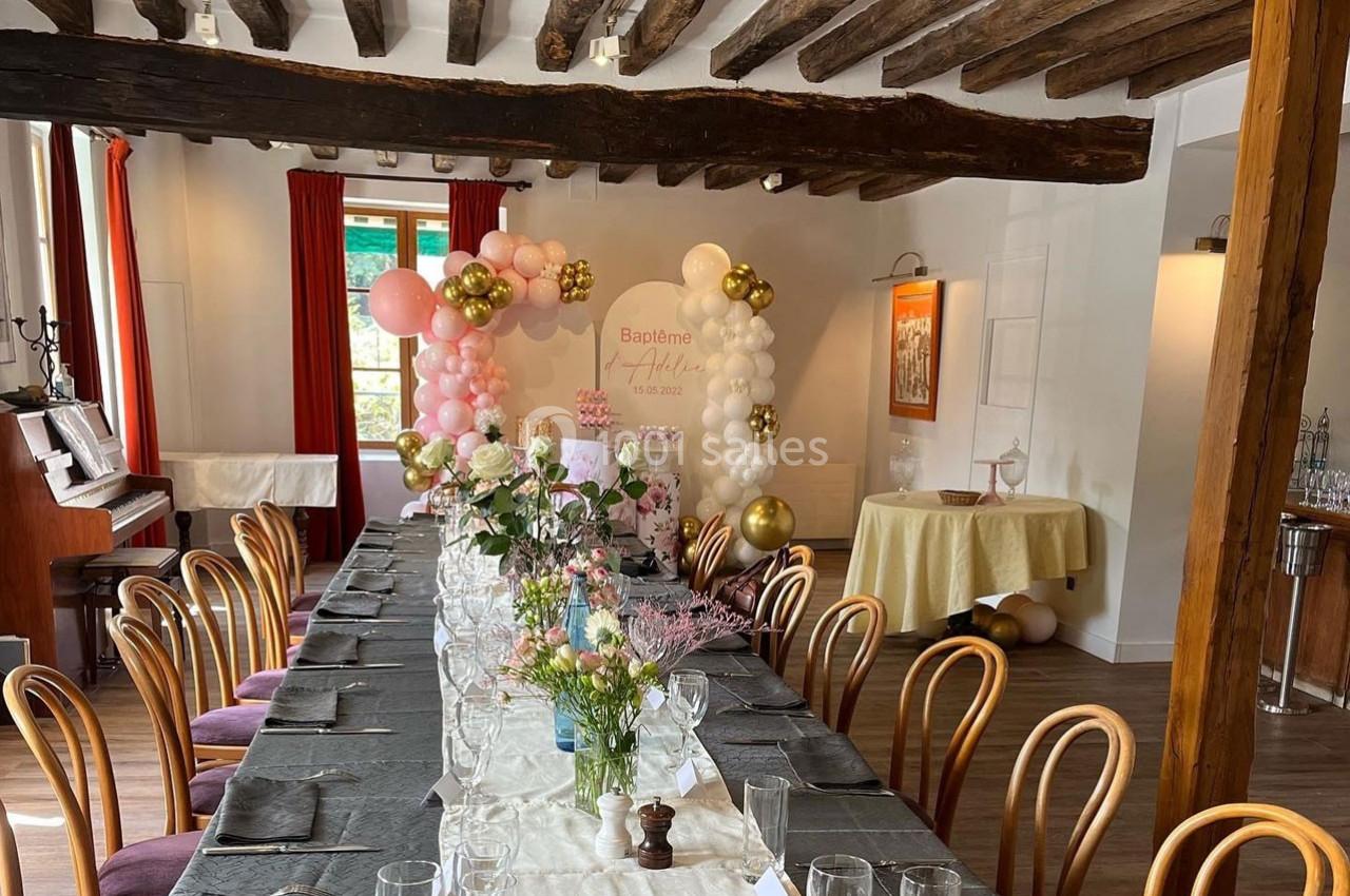 Salle décorée pour une réception avec une longue table dressée, des ballons et des fleurs, sous un plafond à poutres…