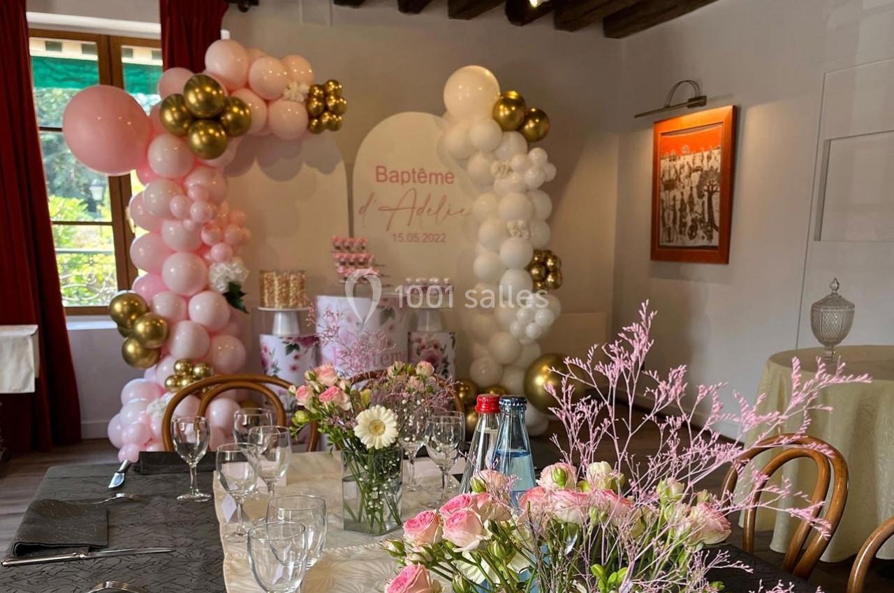 Salle décorée pour un baptême avec ballons roses et blancs, table dressée avec fleurs et vaisselle.