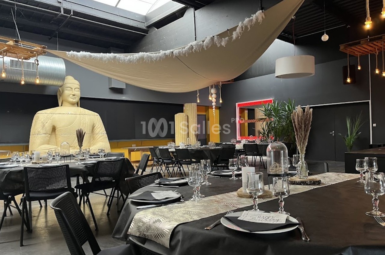 Salle décorée pour un événement avec tables dressées, statue de Bouddha et éclairage tamisé sous une toile suspendue.