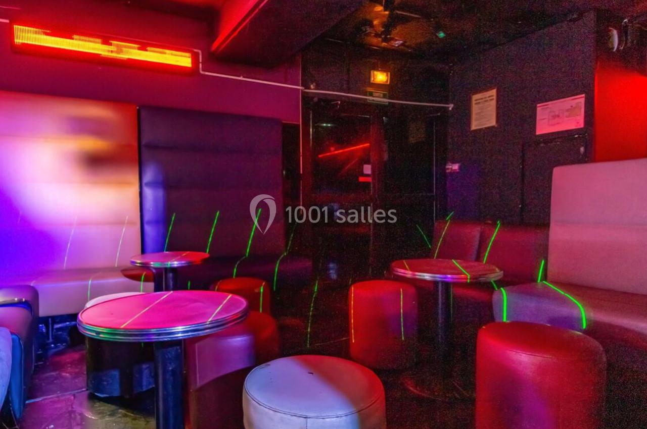 Salle de bar avec banquettes et tables rondes, éclairée par des lumières rouges et néons colorés.