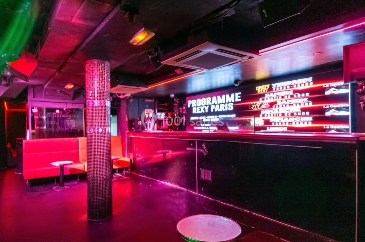 Intérieur d'une boîte de nuit avec banquettes rouges, bar éclairé par des néons et programme affiché sur un écran.