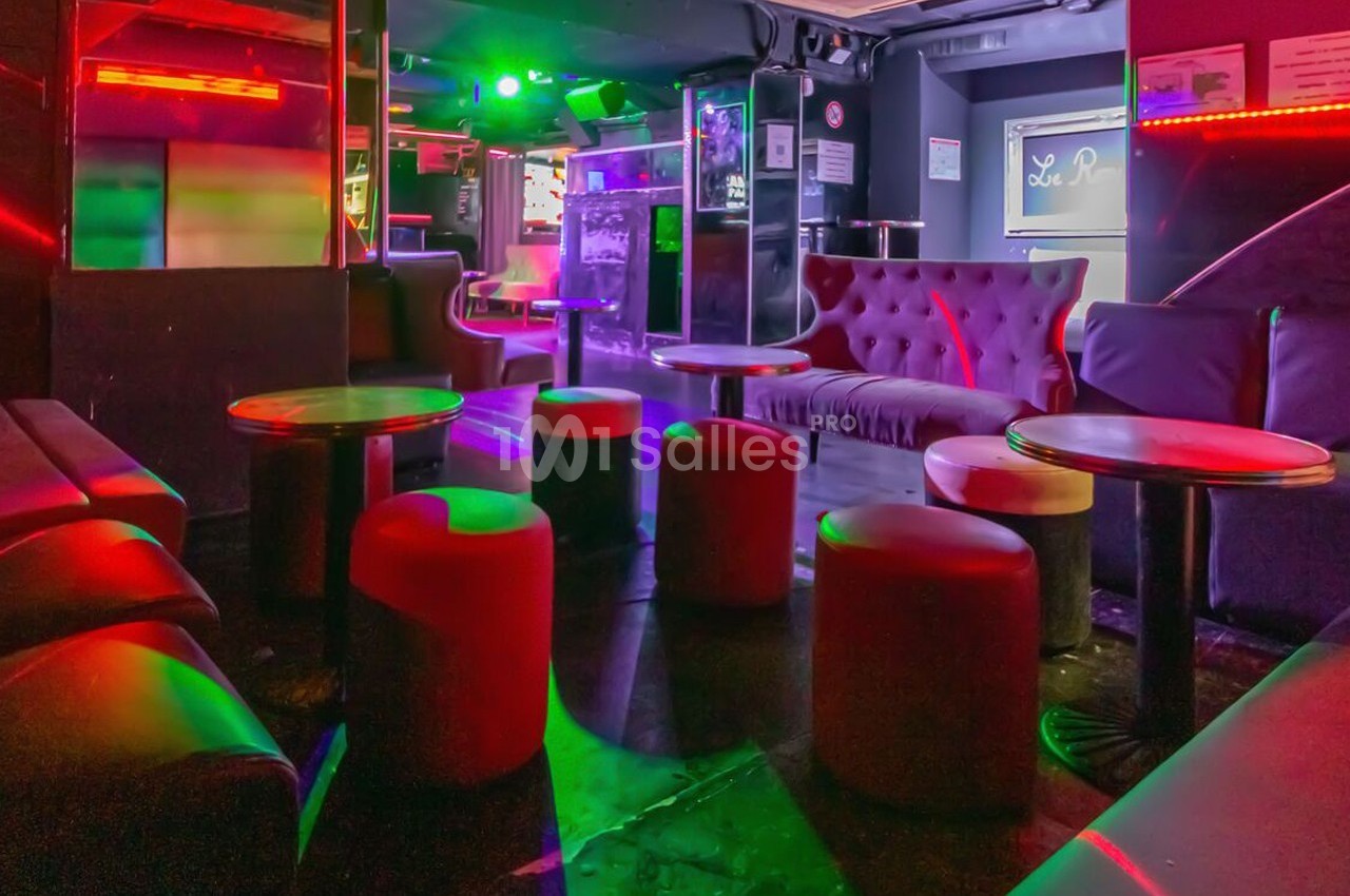 Espace intérieur d'un bar ou club avec canapés, tabourets et tables, éclairé par des lumières colorées.