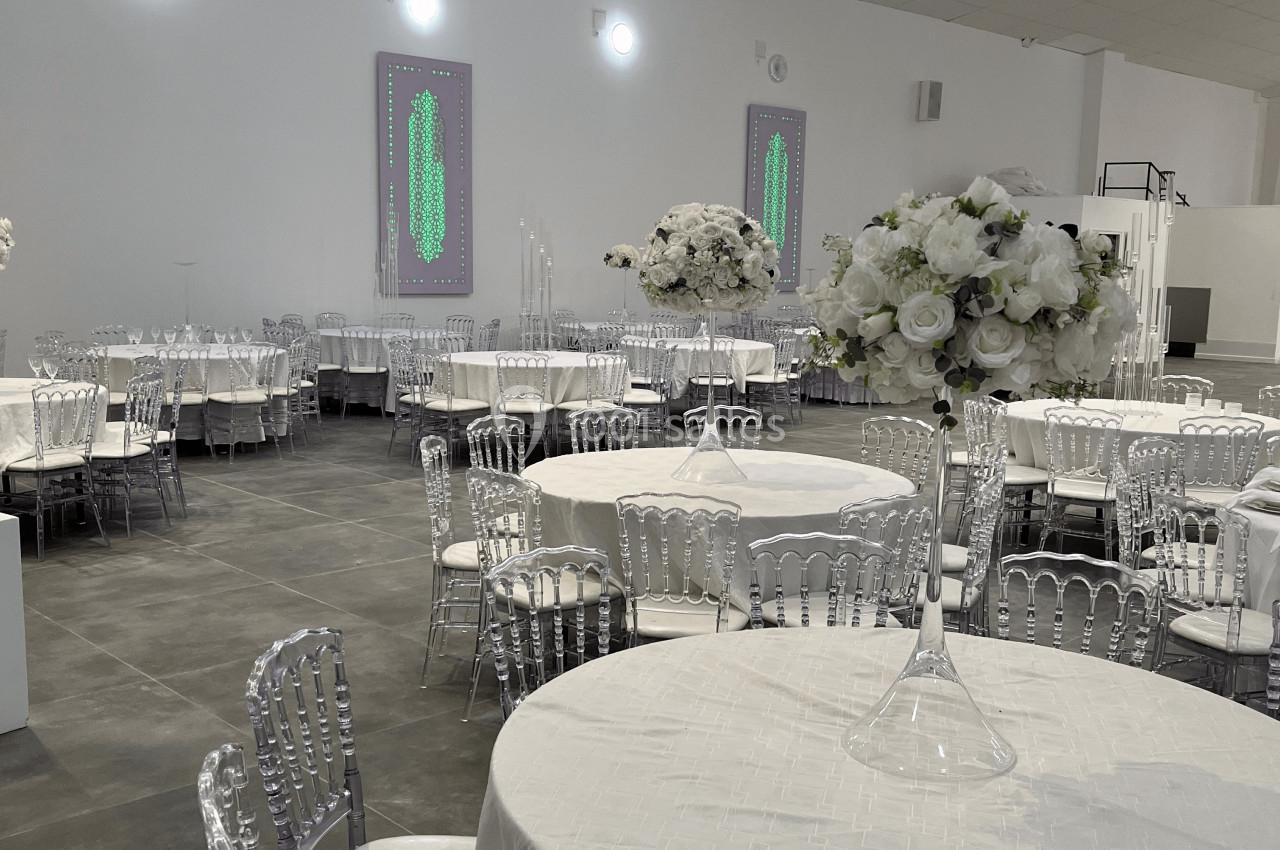 Salle de réception avec tables rondes dressées de nappes blanches, chaises transparentes et décorations florales.