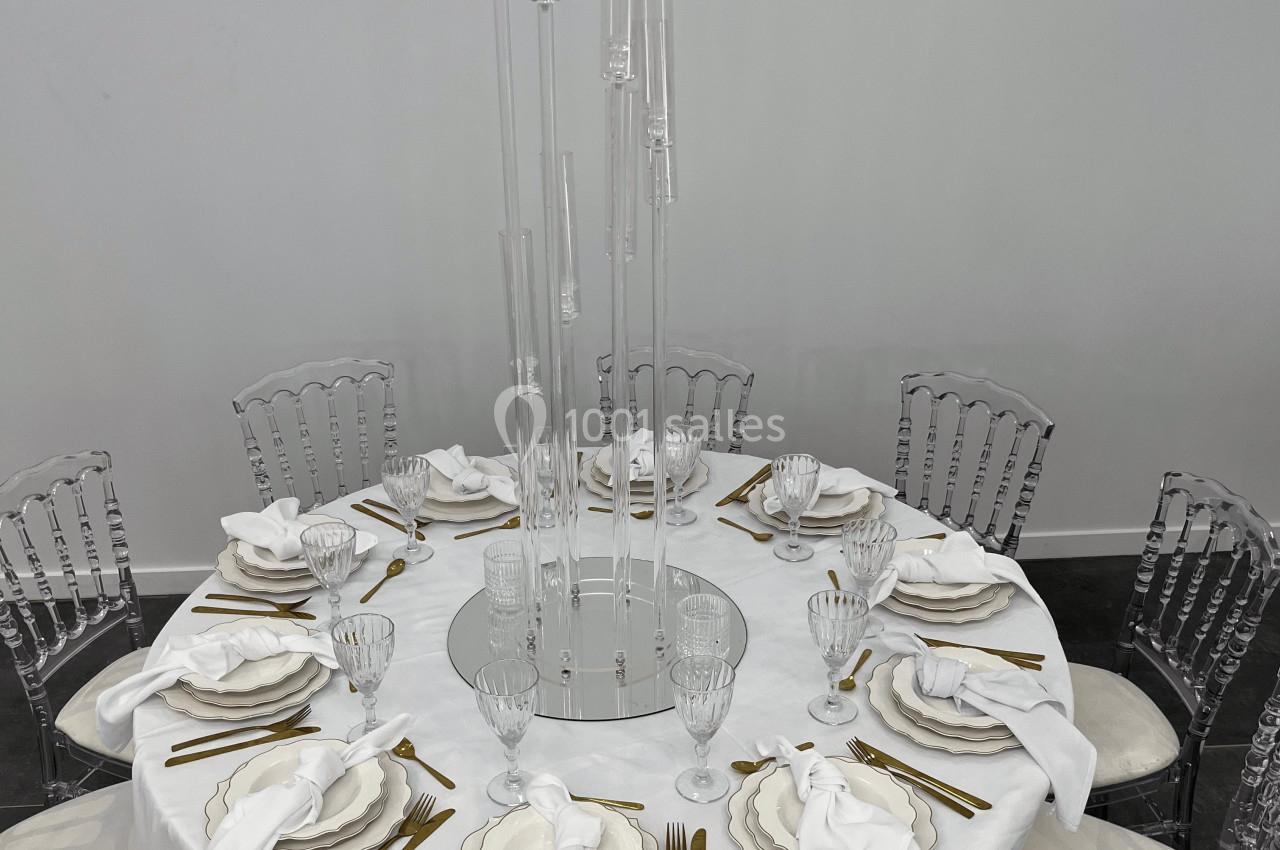Table ronde dressée avec nappe blanche, couverts dorés, serviettes pliées et centre de table en tubes transparents.