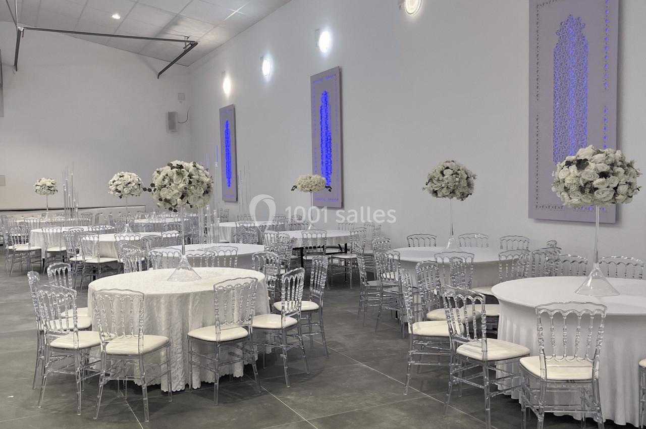 Salle de réception décorée avec des tables rondes, nappes blanches, chaises transparentes et arrangements floraux blancs.