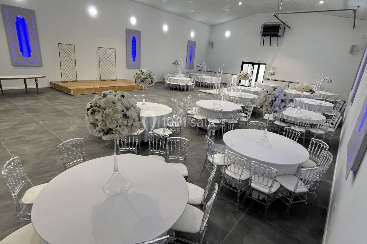 Salle de réception moderne avec tables rondes dressées, chaises transparentes et décorations florales blanches.