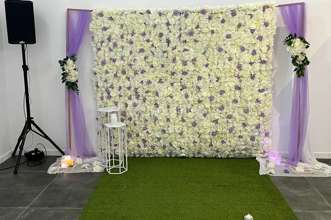 Mur floral blanc et violet décoré de voilages mauves, avec bougies et tabouret blanc sur un tapis vert.