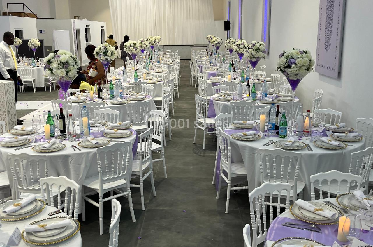 Salle de réception décorée avec des tables rondes, nappes blanches, centres de table fleuris et chaises blanches.