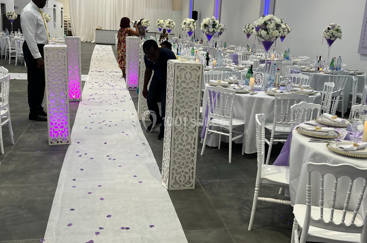 Salle de réception décorée avec des tables dressées, des colonnes ornées et une allée parsemée de pétales violets.