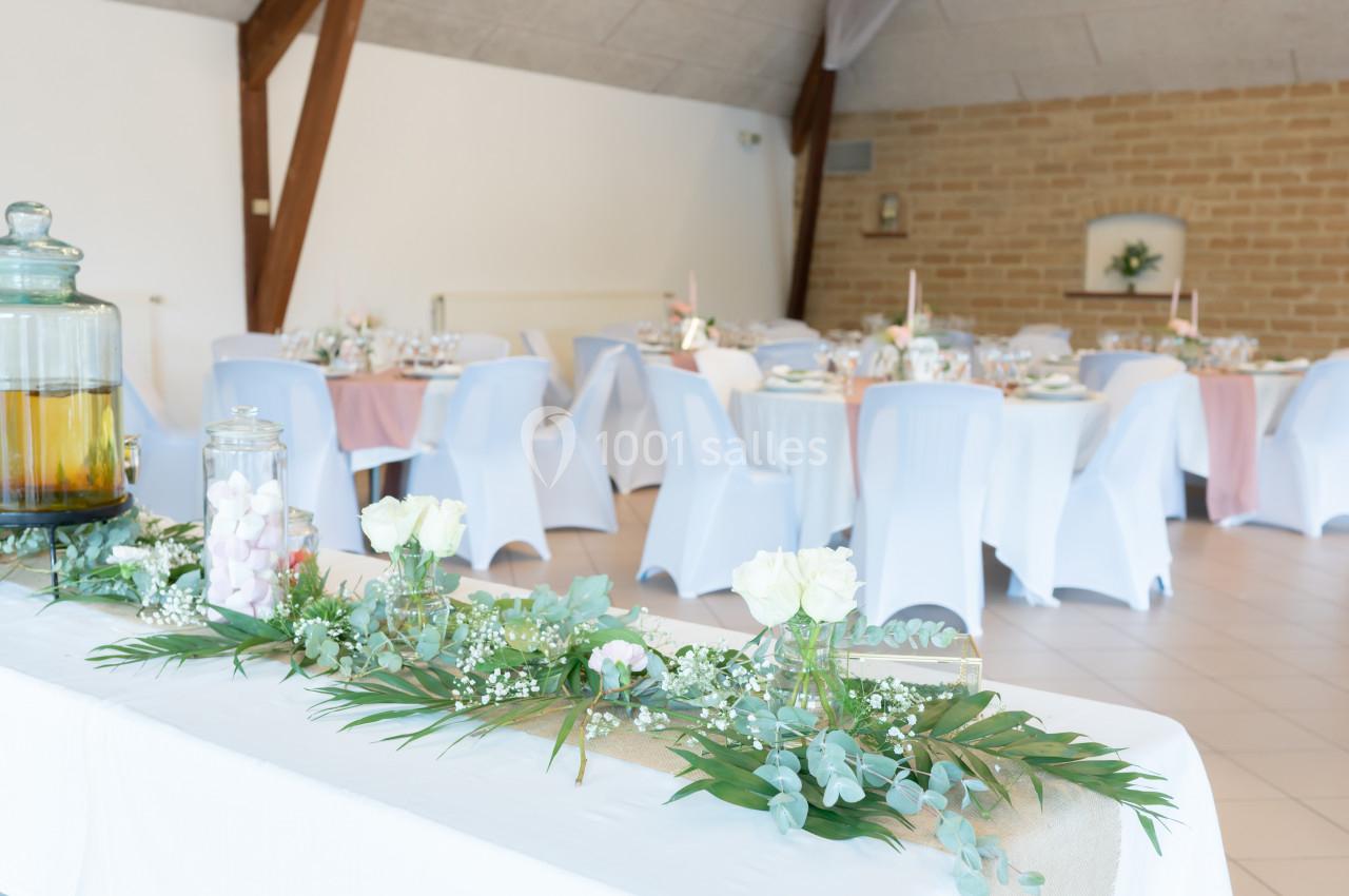 Salle de réception décorée avec des tables dressées, nappes roses et blanches, et un buffet orné de fleurs et feuillages.