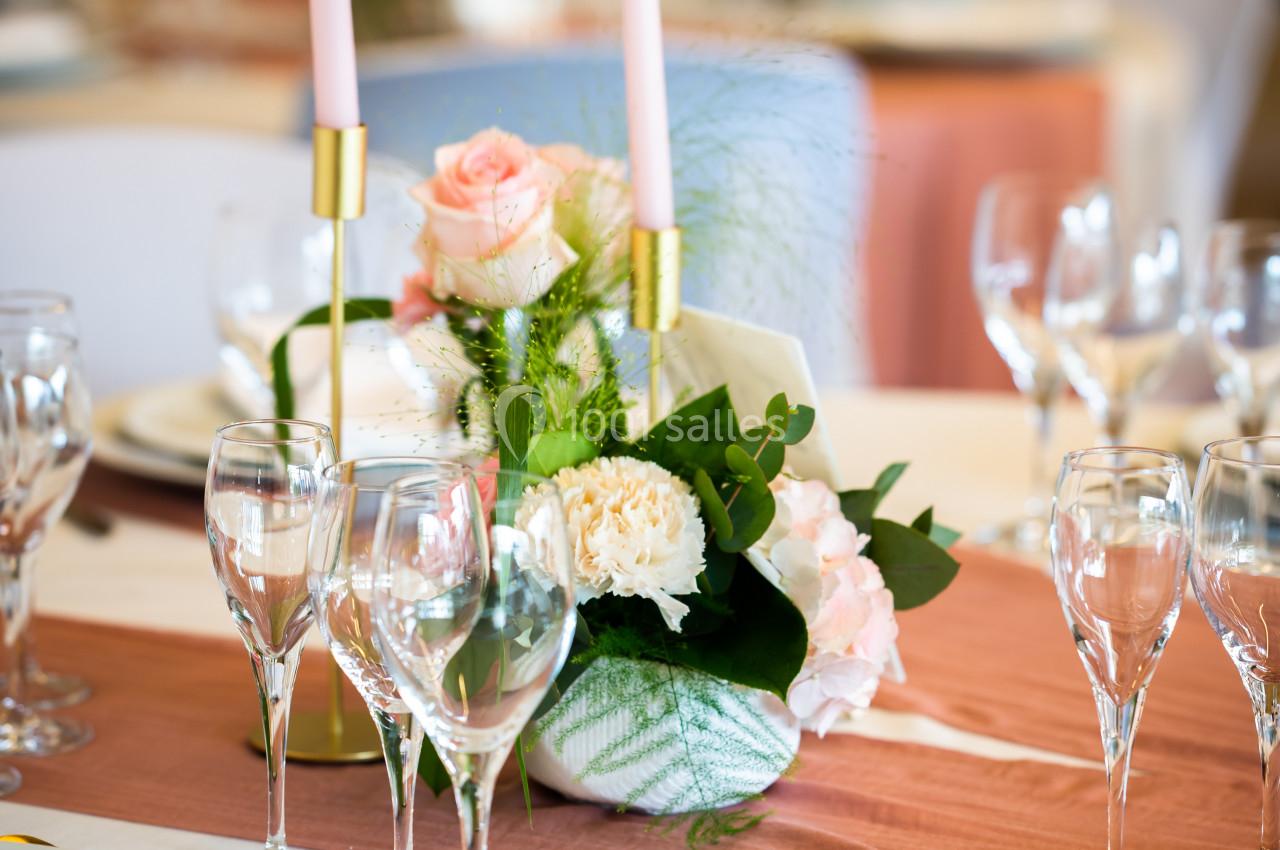 Centre de table floral avec roses et bougies roses sur une nappe blanche, entouré de verres et assiettes.