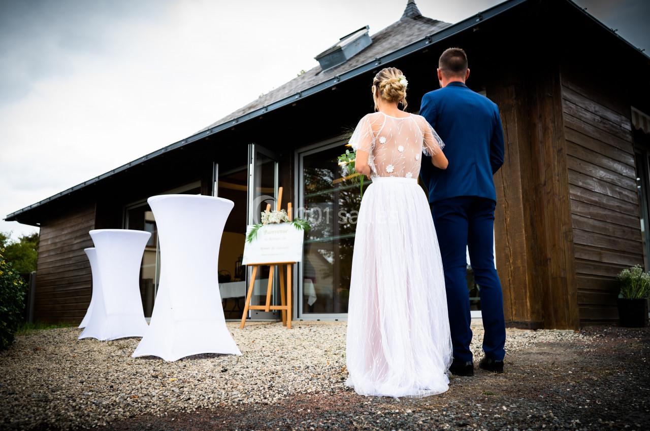 Un couple de dos, en tenue de mariage, se tient devant une salle en bois avec des tables hautes blanches à l'extérieur.