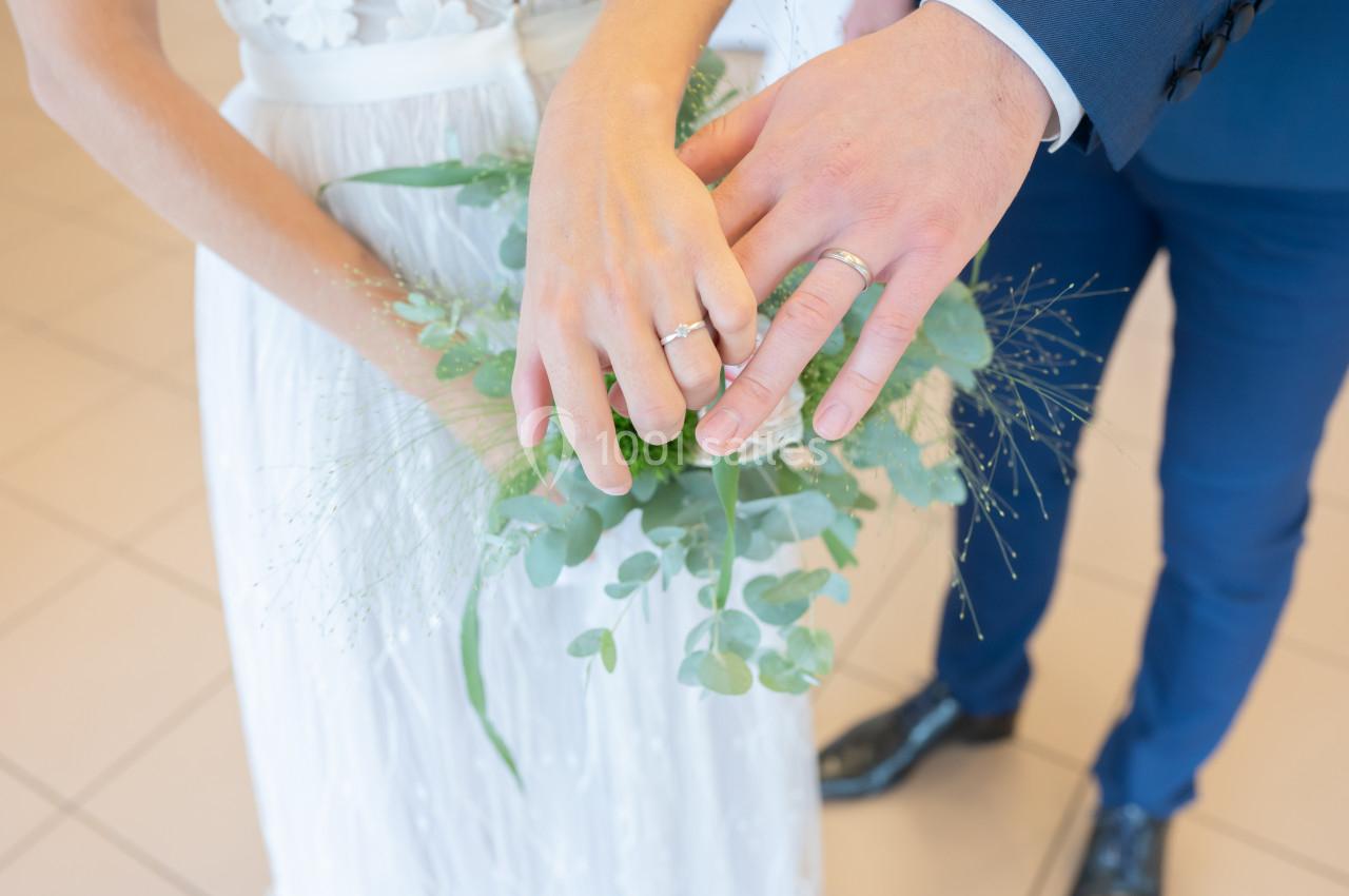 Mains d'un couple en tenue de mariage montrant leurs alliances, avec un bouquet de feuillage en arrière-plan.