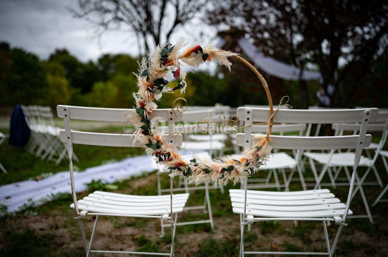Couronne décorative en fleurs séchées suspendue à une chaise blanche dans un cadre extérieur avec des arbres.