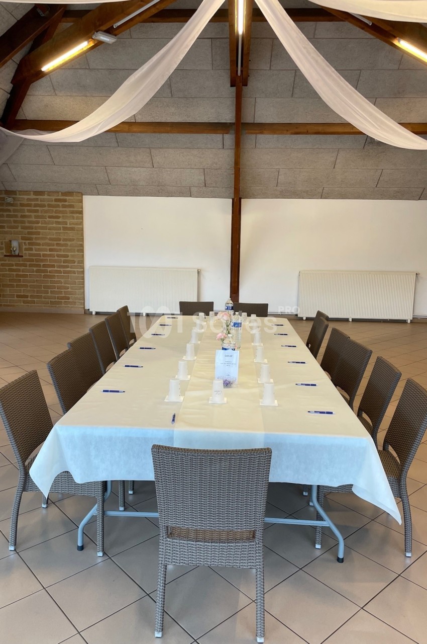Table rectangulaire dressée avec nappes blanches, chaises en osier, stylos et gobelets, dans une salle au plafond en bois.