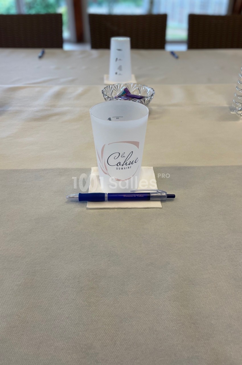 Un gobelet personnalisé, un stylo bleu et une bouteille d'eau disposés sur une table beige dans une salle lumineuse.
