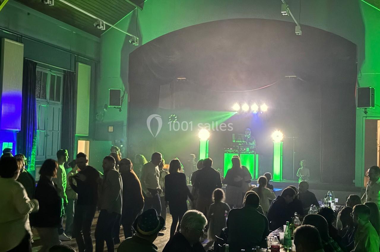 Des personnes dansent et discutent dans une salle éclairée par des lumières vertes, avec un DJ sur une scène au fond.